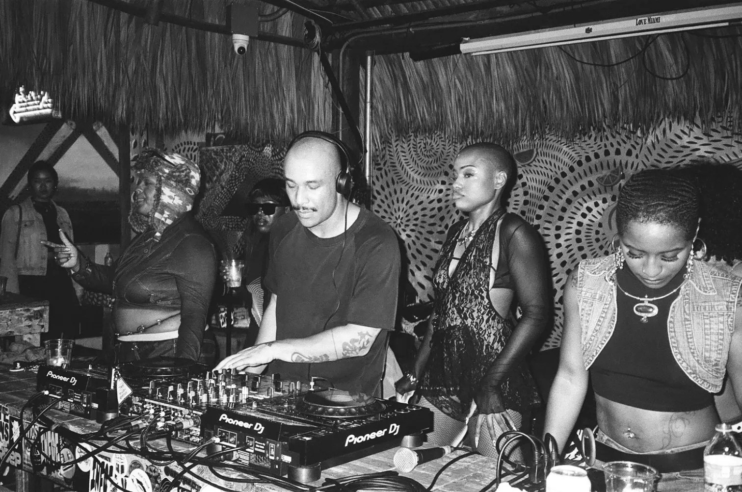 black and white photo of a DJ at the turntables, flanked by several women