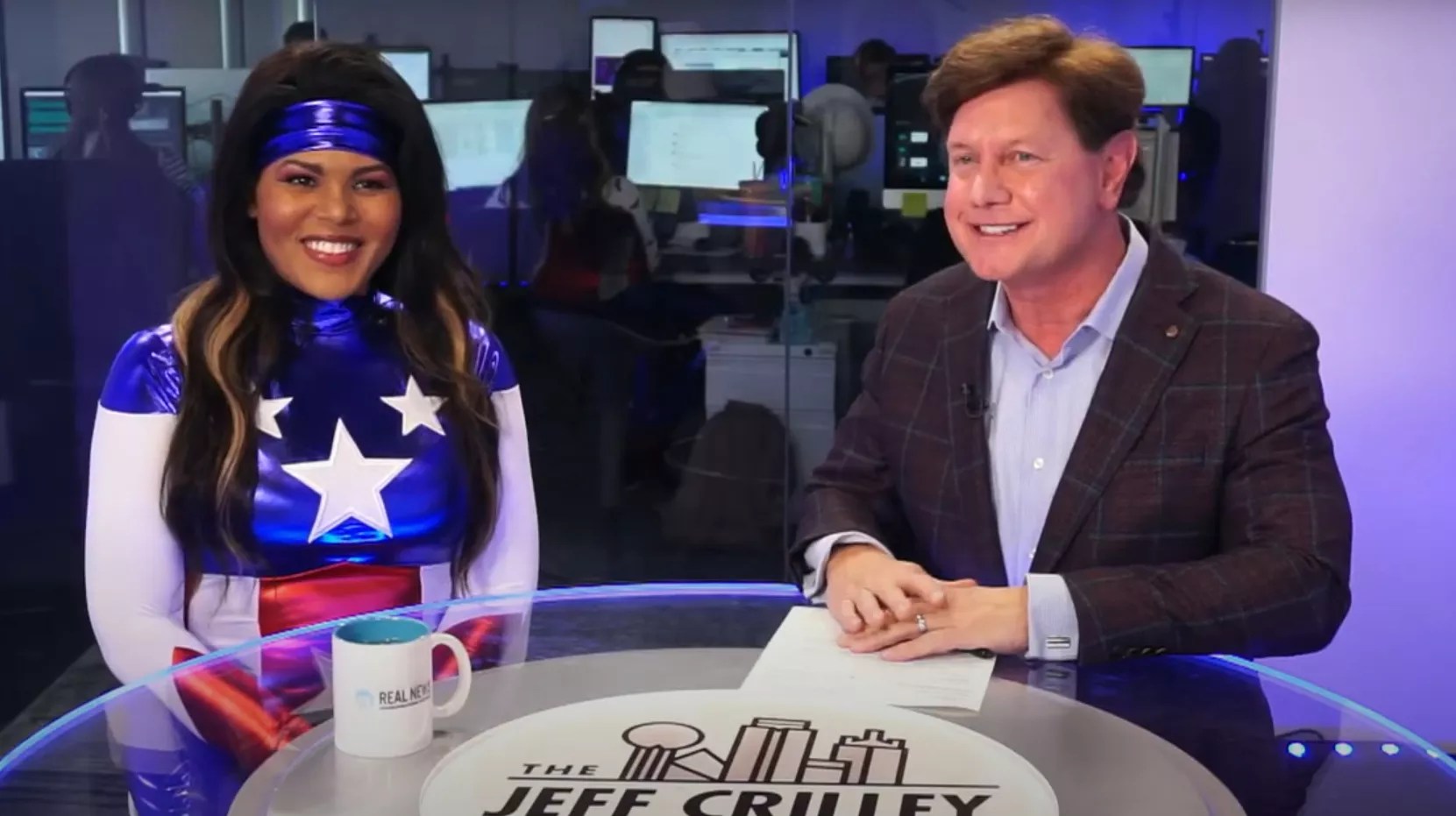 Raven Harrison is wearing a Wonder Woman suit, looking at the camera. She sitting on the left of Jeff Crilley.